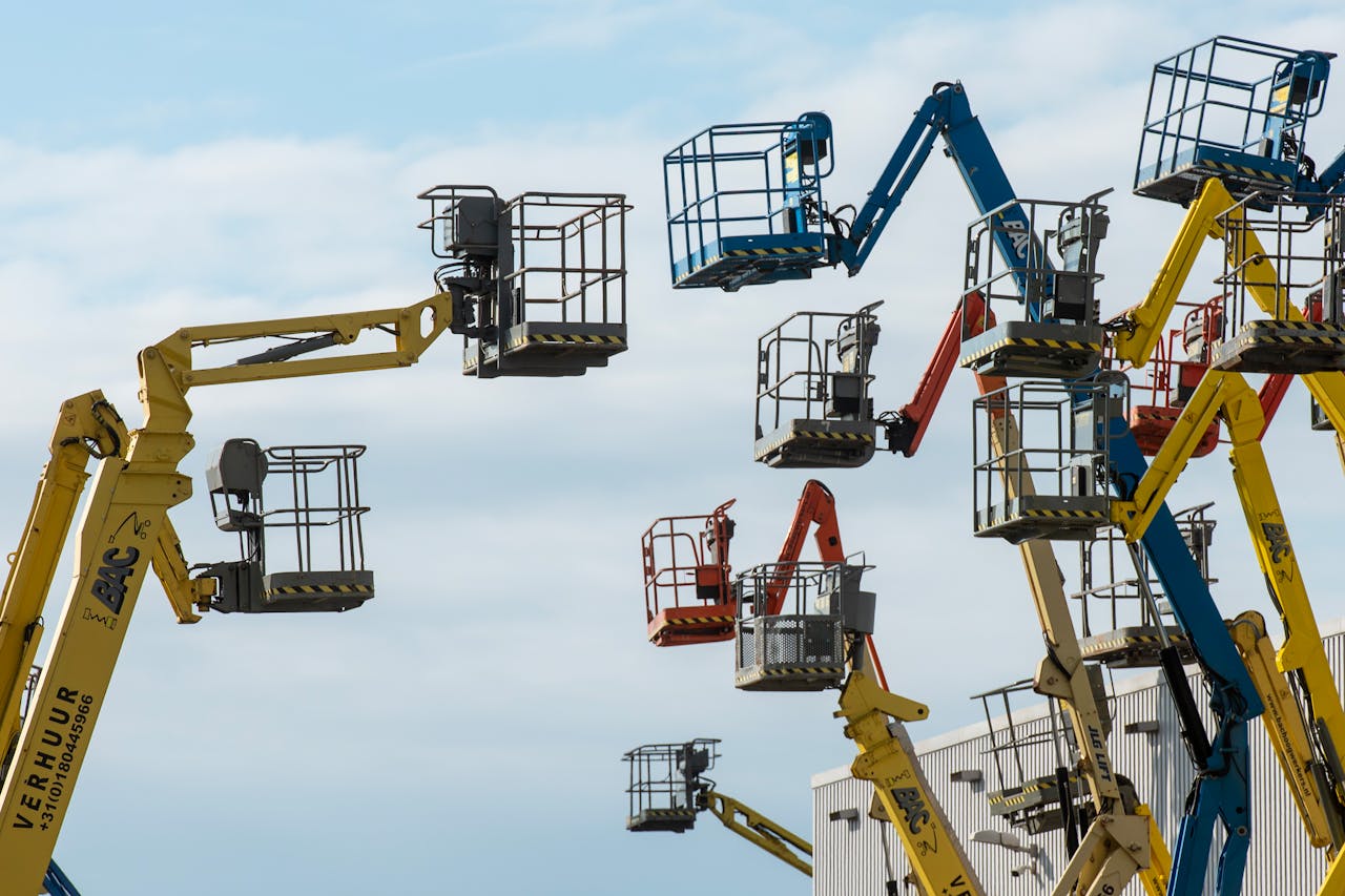 portfolio-01 A collection of colorful boom lifts reaching toward a clear blue sky, showcasing industrial machinery.